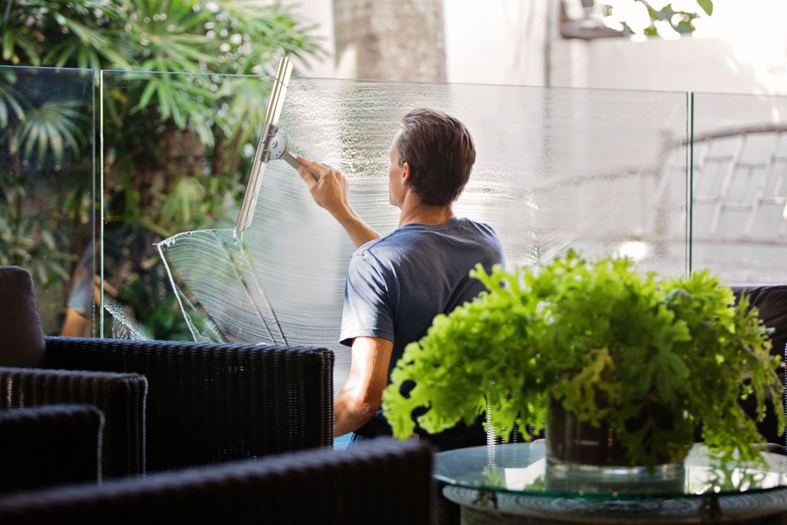 Man using a squeegee on an outdoor glass panel beside garden plants